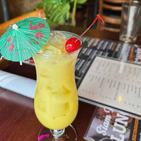 A vibrant yellow cocktail with ice, a red cherry, and a green paper umbrella on a wooden table.