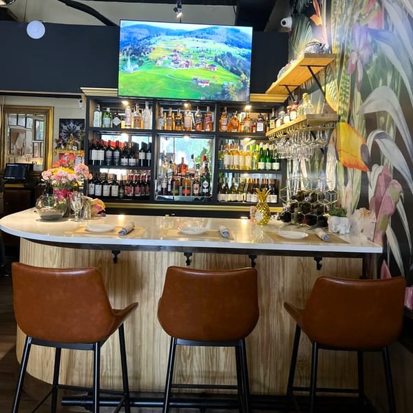 A well-stocked bar at Siam Thai Restaurant with a large TV screen above, and three brown bar stools.