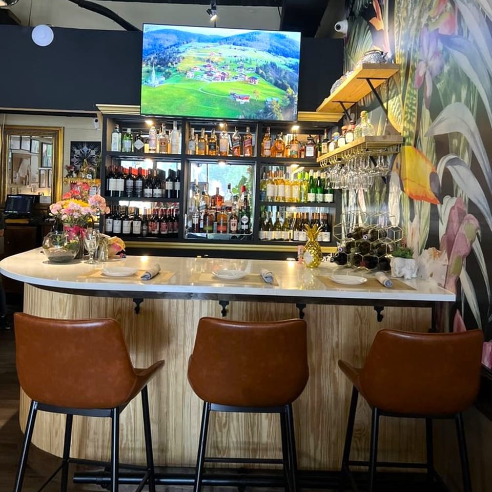 A well-stocked bar at Siam Thai Restaurant with a large TV screen above, and three brown bar stools.